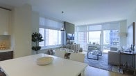 This single-floor space is designed with an open-concept layout that effortlessly connects the dining and living areas, creating a cohesive and functional living environment. Expansive floor-to-ceiling windows wrap around the space, flooding the interiors with abundant natural light while framing striking panoramic views of the surrounding cityscape and waterfront beyond.

The dining area features a sleek white table with matching upholstered chairs, anchored by a contemporary pendant light that adds a refined focal point overhead. Lush potted greenery introduces a natural, organic element that softens the clean, modern aesthetic.

The living area flows seamlessly from the dining space, centered around a plush neutral sofa and a glass coffee table set atop a textured area rug. A wall-mounted flat-screen television is positioned between the windows, while the generous glazing ensures the room remains light-filled from multiple angles throughout the day.

Throughout, the interior palette is kept deliberately neutral — crisp whites, soft greys, and warm natural tones — lending the space a calm, sophisticated ambiance. Roller shades on each window offer flexible light control without compromising the openness of the design. The overall atmosphere is bright, contemporary, and inviting, with the urban skyline serving as a dramatic and ever-present backdrop.