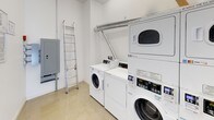 This shared laundry facility is a single-floor, purpose-built space designed for efficient, communal use. The room features a clean, minimal aesthetic with white-painted walls and polished concrete flooring that is easy to maintain and keep tidy.

The space is outfitted with multiple commercial-grade front-loading washers and stacked dryer units, offering ample laundry capacity. The machines are equipped with digital control panels and accept card-based payments, providing a modern and convenient laundry experience. Ventilation ducting runs overhead, connecting the dryers to the building's exhaust system for safe and effective operation.

An electrical panel is mounted on the wall and is easily accessible, alongside a utility ladder for maintenance purposes. A black metal utility pipe runs vertically along one wall, indicating the room's integration with the building's infrastructure. Safety is addressed through the presence of a fire extinguisher and a fire alarm mounted near the exit door.

An information board displaying laundry instructions and service contact details is posted on the wall, offering guidance for users. A waste bin is conveniently positioned near the machines. The open doorway connects the laundry room to an adjacent well-lit corridor, ensuring easy access. Overall, the space is organized, functional, and well-equipped to serve the needs of building residents.