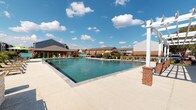 This community amenity space combines a fully equipped fitness center with a spacious outdoor swimming pool, offering residents a comprehensive recreational experience. The fitness center is designed with vaulted ceilings accented by exposed natural wood beams and a deep teal ceiling finish, creating a striking architectural character. Large windows line the walls, flooding the interior with natural light while providing open views of the surrounding community grounds. The gym is well-stocked with a wide range of equipment, including cable machines, weight benches, a full dumbbell rack, elliptical trainers, treadmills, and stationary bikes. Full-length mirrors along one wall enhance the sense of space and functionality. A tiled accent wall near the entryway features a dual-level water fountain station and bottle-filling unit, adding a practical and polished touch. Double glass doors with wood-framed trim connect the spaces cleanly. The outdoor pool area is equally impressive, featuring a generously sized rectangular swimming pool with clear blue water, a surrounding concrete deck, and a white pergola structure anchored by brick columns. Lounge chairs, umbrellas, and landscaped borders complete the inviting outdoor setting beneath open skies.