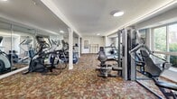 Located at 2607 Welsh Rd in Philadelphia, PA, this fitness center offers a thoughtfully designed single-floor exercise space suited for a full range of workout routines. The room is carpeted in a warm, patterned floor covering and features white-painted structural columns that define distinct workout zones without enclosing the space. Large windows along one wall invite natural light and provide views of the surrounding greenery, creating an open and energizing atmosphere. Recessed and flush-mounted ceiling lights ensure consistent illumination throughout.

The cardio zone includes multiple treadmills, elliptical cross-trainers, and a stationary bike, arranged to allow comfortable use. A full-length mirrored wall expands the sense of space and allows users to monitor their form during exercise. The strength training area is equipped with a multi-station cable machine and a dedicated free-weight section featuring medicine balls, stability balls, resistance bands with a TRX suspension system, and a rack of accessories. A wall-mounted flat-screen television adds convenience for those looking to stay entertained during workouts. Storage cabinetry is also present, contributing to the room's organized layout. The overall design balances functionality and comfort, making this fitness center a practical and well-appointed amenity.