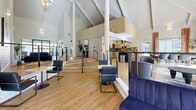 Located at 14800 NW Cornell Rd in Portland, Oregon, this single-floor clubhouse is an architecturally distinctive gathering space defined by its dramatic vaulted ceiling with exposed white-painted beams that rise to impressive heights, creating an airy and welcoming atmosphere throughout. Light hardwood flooring runs continuously across the open layout, tying together the various seating and activity zones seamlessly.

The space is organized into several functional areas across a single, flowing floor plan. A central lounge area features a sculptural navy velvet sofa set against a gallery wall with curated abstract artwork, flanked by minimalist side tables. Adjacent to this is a sunlit multi-seat lounge with low tables and sleek black chairs arranged at split levels, connected by short staircases that add architectural interest.

A dedicated dining area accommodates a large dark-wood table with seating for eight, bathed in natural light pouring through floor-to-ceiling windows draped in warm terracotta linen curtains. These expansive windows frame panoramic views of the surrounding green lawn and residential buildings beyond. A fully equipped kitchen with dark stone countertops and modern stainless steel appliances serves the space at the rear.

Wall-mounted sconces and a statement chandelier provide warm ambient lighting, complementing the abundant natural light. The overall ambiance is modern, refined, and inviting — ideal for communal gatherings and events.