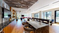 This thoughtfully designed amenity lounge is organized into several distinct zones, each offering its own character while maintaining a cohesive modern aesthetic throughout. Warm hardwood flooring flows continuously across the entire space, grounding the varied design elements and unifying the layout.

The centerpiece of the lounge is a dramatic oversized island clad in dark, veined marble, paired with contemporary swivel bar stools. Above, a reclaimed wood ceiling panel adds rustic contrast to the otherwise clean, modern finishes, while a sculptural branching chandelier provides an elegant focal point. Adjacent to the island, warm wood cabinetry with open display niches lines the wall, offering both storage and visual interest alongside a long dark-countered bar area.

The lounge transitions into a comfortable seating area defined by bold crimson sofas and navy accent chairs arranged around a low coffee table atop a graphic patterned rug. A wall-mounted flat-screen television and a marble-surround fireplace anchor this zone, creating an inviting gathering spot. A coffered circular ceiling detail above adds architectural dimension.

Large floor-to-ceiling windows and generously sized panes throughout flood the space with natural light and frame views of the surrounding outdoor areas, enhancing the open, welcoming ambiance of this well-appointed communal lounge.