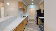 This single-floor 1-bedroom, 1-bathroom unit offers a clean, modern aesthetic throughout. The open living area is finished with durable wood-look plank flooring in a cool gray tone, neutral beige walls, and recessed lighting that creates a bright and welcoming atmosphere. A baseboard heater and window with blinds are also present near the entryway, which features a solid white panel door with a modern lever handle. The galley-style kitchen is efficiently designed with warm honey-toned cabinetry, crisp white countertops, a stainless steel sink, and black appliances including a refrigerator, electric range, and built-in microwave. Recessed lighting overhead keeps the space well-lit. The bedroom is carpeted for comfort and includes a glass-paneled exterior door with blinds, allowing for natural light while maintaining privacy. A standard interior door connects to the rest of the unit. The bathroom is outfitted with a tub and shower combination enclosed by sliding glass doors, a toilet, a wall-mounted vanity mirror, and a single vanity with wood-toned cabinetry. Consistent wood-look flooring carries into the bathroom, tying the unit's design together cohesively. Overall, the unit presents a clean, move-in-ready space with functional, modern finishes throughout.