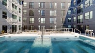 This contemporary residential building showcases a striking industrial-modern exterior clad in dark and light metal paneling, with private balconies extending from multiple floors. At ground level, a sparkling outdoor pool serves as a central amenity, framed by clean white railings and decorative string lighting that adds a welcoming ambiance.

The interior amenity spaces are thoughtfully designed with a cohesive sage-green color palette and light hardwood-style flooring throughout. The lounge area offers generous seating with comfortable sectional sofas, large windows that flood the space with natural light, and direct access to an outdoor terrace. A separate media lounge provides a more intimate gathering space, featuring a large wall-mounted television, built-in dark wood cabinetry with gold hardware, and warm sconce lighting.

The amenity kitchen is a standout feature, boasting a dramatic black marble-inspired backsplash, sleek grey cabinetry, white quartz countertops, and exposed industrial ductwork overhead. Pendant globe lighting adds a polished finishing touch above the spacious island with bar seating.

The rooftop terrace is an expansive outdoor retreat with wide-open views of the surrounding cityscape and distant mountain ranges. Raised planters with desert landscaping, seating areas, and open concrete decking make it an ideal space for relaxing or socializing under open skies.
