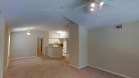 This single-floor residence offers a thoughtfully designed open-concept layout that seamlessly connects the living area to the kitchen, creating a spacious and functional flow throughout the home. The living area is generously sized, featuring soft wall-to-wall carpeting in warm neutral tones that contribute to a cozy and inviting ambiance. A ceiling fan with integrated lighting is centrally positioned, providing both comfort and illumination to the space.

The kitchen is well-appointed and anchored by a curved breakfast bar that serves as a natural dividing element between the cooking and living areas. White cabinetry lines the upper and lower sections of the kitchen, complemented by white appliances including a refrigerator and range with overhead ventilation. The kitchen benefits from its own dedicated overhead lighting, keeping the workspace bright and functional.

The walls throughout are finished in a clean, neutral beige palette, lending a calm and cohesive aesthetic to the interior. A hallway visible from the main living area suggests additional rooms beyond, indicating a practical and well-organized floor plan. White baseboards and door trim provide a crisp contrast to the warm carpet and wall tones, adding a polished finishing detail to the overall design.
