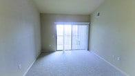This single-floor space features a straightforward, functional layout designed to maximize comfort and usability. The main living area is finished with wall-to-wall carpeting in a neutral tone, providing a soft, cohesive foundation that complements the surrounding beige walls. A sliding glass door serves as a prominent architectural feature, drawing in abundant natural light and providing direct access to a private balcony, making the interior feel open and connected to the outdoors. Vertical blinds accompany the sliding door, allowing for adjustable light control and privacy. The ceiling is standard height and includes a centrally placed HVAC vent, ensuring efficient climate control throughout the space. Clean white baseboards frame the perimeter of the room, adding a crisp contrast to the neutral walls and carpet. The overall ambiance is calm and understated, offering a blank canvas well-suited for a variety of interior arrangements. The space is move-in ready, with its simple, clean aesthetic providing flexibility for personalization.