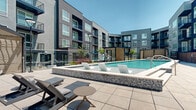 This contemporary multi-story residential complex showcases a striking modern architectural design, characterized by clean lines, bold dark and light contrasting exterior facades, large windows, and private balconies spanning multiple levels. The outdoor amenity area serves as the centerpiece of the community, offering a well-appointed resort-style experience. A sparkling swimming pool, accented by elegant mosaic tile detailing and stainless steel entry railings, is complemented by a shallow sun shelf fitted with sleek lounge chairs—ideal for relaxation. Adjacent to the pool, a dedicated spa area and stylish shaded cabanas with comfortable seating provide additional retreat options. The surrounding pool deck features generous tiled patio space and rows of lounge chairs for sunbathing. A separate landscaped courtyard area offers a more tranquil setting, with raised concrete planters filled with ornamental grasses and native plantings, bistro-style seating, and open patio space. Recreational amenities such as a cornhole game area add a social element to the outdoor environment. Thoughtfully designed ramps and railings ensure accessibility throughout. The entire outdoor space is framed by the multi-floor building, creating an enclosed, community-oriented atmosphere bathed in natural sunlight.