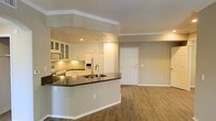 This single-floor space showcases an open-concept layout that seamlessly connects the kitchen and living area, creating a cohesive and functional environment. The kitchen is outfitted with white cabinetry, including glass-front upper cabinets that add a refined touch, complemented by a mosaic tile backsplash and dark countertops that provide a striking contrast. A built-in microwave and white refrigerator with an ice and water dispenser complete the appliance suite. The peninsula, featuring a double-basin sink with a modern faucet, extends the counter space while serving as a natural divider between the kitchen and the open living area. Crown molding runs throughout the ceiling, adding a polished architectural detail, while recessed lighting in the kitchen ensures ample illumination. The living area is anchored by warm hardwood-style flooring that extends throughout, contributing to a cohesive and inviting atmosphere. Neutral-toned walls provide a versatile backdrop. A paneled entry door with standard hardware is visible, offering a clean and classic aesthetic. The overall ambiance is bright, modern, and well-appointed.