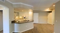 This single-floor space showcases an open-concept layout that seamlessly connects the kitchen and living area, creating a cohesive and functional environment. The kitchen is outfitted with white cabinetry, including glass-front upper cabinets that add a refined touch, complemented by a mosaic tile backsplash and dark countertops that provide a striking contrast. A built-in microwave and white refrigerator with an ice and water dispenser complete the appliance suite. The peninsula, featuring a double-basin sink with a modern faucet, extends the counter space while serving as a natural divider between the kitchen and the open living area. Crown molding runs throughout the ceiling, adding a polished architectural detail, while recessed lighting in the kitchen ensures ample illumination. The living area is anchored by warm hardwood-style flooring that extends throughout, contributing to a cohesive and inviting atmosphere. Neutral-toned walls provide a versatile backdrop. A paneled entry door with standard hardware is visible, offering a clean and classic aesthetic. The overall ambiance is bright, modern, and well-appointed.