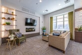 The Tenant Lounge at 39 Florence St in Malden, MA is a thoughtfully designed communal amenity space that offers residents a sophisticated and comfortable environment for relaxing or socializing. The lounge is organized into distinct functional zones that flow seamlessly together.

The main seating area centers around a large wall-mounted flat-screen television paired with a sleek linear fireplace set against a textured tile accent wall. A generous sectional sofa in warm golden tones anchors the space, complemented by coordinating accent chairs and decorative greenery. Natural light pours in through oversized windows dressed with olive-toned drapes, offering views of the surrounding neighborhood.

A dedicated dining nook features high-top tables and upholstered bar-height chairs arranged beneath another wall-mounted television, with large windows on multiple walls creating a bright, airy atmosphere. A cozy reading corner provides an intimate retreat, complete with a leather wingback chair and illuminated floating wood shelves set against a white tile backdrop.

The kitchenette is well-appointed with rich walnut cabinetry, white quartz countertops, a stainless steel sink, an under-counter beverage refrigerator, a microwave, and an eye-catching mosaic stone tile backsplash. Warm hardwood-style flooring in the kitchen transitions to patterned carpet tiles throughout the lounge, tying the space together with a modern, welcoming aesthetic.