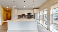 This single-floor residence showcases a thoughtfully designed open-concept layout with contemporary finishes throughout. The kitchen serves as a central highlight, featuring flat-panel white cabinetry extending to the ceiling, a rich dark brick-style tile backsplash, and a full suite of stainless steel appliances including a gas range, built-in microwave, and French door refrigerator. A generously sized white waterfall island anchors the space, with cylindrical pendant lights suspended above providing focused task lighting alongside recessed ceiling fixtures. Large floor-to-ceiling windows along the kitchen's perimeter flood the area with natural light and offer outdoor views.

The living area continues the light aesthetic with wood-look flooring and direct access to a private balcony through wide windows. The bedroom features plush carpeting and a large picture window framing a scenic waterfront and cityscape view.

Two bathrooms are finished cleanly in neutral tones with wood-look flooring. One offers a soaking tub with full tile surround, while the other features a walk-in glass-enclosed shower and a long vanity with ample storage. The entryway is functional and well-proportioned, completed with a modern door and keypad entry.