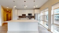 This single-floor residence showcases a thoughtfully designed open-concept layout with contemporary finishes throughout. The kitchen serves as a central highlight, featuring flat-panel white cabinetry extending to the ceiling, a rich dark brick-style tile backsplash, and a full suite of stainless steel appliances including a gas range, built-in microwave, and French door refrigerator. A generously sized white waterfall island anchors the space, with cylindrical pendant lights suspended above providing focused task lighting alongside recessed ceiling fixtures. Large floor-to-ceiling windows along the kitchen's perimeter flood the area with natural light and offer outdoor views.

The living area continues the light aesthetic with wood-look flooring and direct access to a private balcony through wide windows. The bedroom features plush carpeting and a large picture window framing a scenic waterfront and cityscape view.

Two bathrooms are finished cleanly in neutral tones with wood-look flooring. One offers a soaking tub with full tile surround, while the other features a walk-in glass-enclosed shower and a long vanity with ample storage. The entryway is functional and well-proportioned, completed with a modern door and keypad entry.