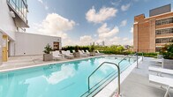 This exceptional pool amenity offers two distinct aquatic experiences designed for relaxation and recreation. The expansive outdoor pool takes center stage with a generously sized swimming area surrounded by light-toned tile decking and lined with rows of white lounge chairs and potted greenery. The elevated rooftop setting provides unobstructed panoramic views of the Atlanta city skyline, framed by a lush hedgerow border and open skies — making it a standout feature for outdoor leisure. The adjacent indoor pool provides a more intimate and sheltered alternative, enclosed within a thoughtfully designed space featuring floor-to-ceiling vertical wood paneling and a warm, natural wood-clad ceiling. Circular porthole-style mirrors accent the walls, adding an architectural touch that complements the spa-like atmosphere. A comfortable lounge area with upholstered seating is integrated directly into the indoor pool space, creating an inviting environment for relaxation year-round. The surrounding building architecture blends modern white paneling with warm wood accents and large glass doors, maintaining a cohesive design language between the indoor and outdoor areas. Together, these spaces offer a well-appointed, resort-caliber pool experience with a strong urban aesthetic.