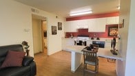 This single-floor space offers a functional and straightforward layout with an open-concept kitchen and dining area as its central feature. The kitchen is equipped with white cabinetry that extends to the upper walls, providing ample storage space. A bold red accent wall serves as a striking visual focal point, adding warmth and personality to the area. The kitchen includes a white electric range with an overhead exhaust hood, a black oven, and a cooktop, making it a practical space for everyday cooking. A compact kitchen island doubles as a dining table, accompanied by simple wooden chairs, creating an efficient eat-in dining setup. The flooring throughout is a warm wood-look laminate that contributes to a cohesive and inviting atmosphere. A hallway visible from the kitchen leads to additional rooms, including what appears to be a bathroom with tiled flooring, suggesting a well-connected layout. Recessed fluorescent lighting in the kitchen ensures bright, even illumination throughout the space. The overall design is practical and low-maintenance, suited for comfortable everyday living.