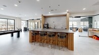 Located at 9 John St in Clinton, Connecticut, this single-floor amenity space offers a thoughtfully designed, open-plan layout that blends social, recreational, and functional areas seamlessly. The interior is defined by a cohesive modern aesthetic, featuring large-format light stone tile flooring, crisp white ceilings with recessed lighting, and striking vertical wood slat wall panels that add warmth and texture throughout.

At the heart of the space is a central kitchen island wrapped in slatted wood paneling and topped with a dark stone countertop, surrounded by upholstered bar stools with brass-toned legs. The kitchen area includes a sink, coffee station, and a tiled stone backsplash, creating a polished yet welcoming gathering point.

Adjacent to the kitchen, a recreation zone features a full-size pool table alongside floor-to-ceiling glass doors that open to an outdoor terrace, allowing abundant natural light to fill the space. A dedicated lounge area provides comfortable seating arranged around a wall-mounted display screen, ideal for informal meetings or socializing.

A collaborative workspace area includes a built-in desk station positioned beneath large windows overlooking the streetscape. Glass-partitioned rooms offer additional private or semi-private areas within the open layout. The entry welcomes visitors with a branded floor mat and Adirondack-style seating positioned beside expansive black-framed glass doors.