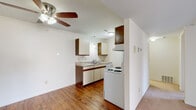 This single-floor residence presents a practical and straightforward layout across its living spaces. The kitchen is a functional galley-style space equipped with white appliances, including an electric coil range, dishwasher, and full-size refrigerator. Cabinetry features cream-colored lower cabinets accented with dark wood trim, complemented by matching upper cabinets, granite-patterned countertops, a double-basin sink, and a pendant light. Hardwood flooring runs through the kitchen and extends into adjacent areas.

The living area is carpeted and bright, featuring a ceiling fan with integrated lighting and a sliding glass door that allows natural light and provides views of an outdoor green space. A secondary room features hardwood flooring, a built-in closet with bifold doors, and direct access to the main living space.

The bedrooms are carpeted and neutral in tone, each featuring windows with horizontal blinds that allow natural light. The bathroom includes a full soaking tub and shower combination with sage-green square wall tile, stainless steel grab bars for accessibility, and hardwood flooring. The overall aesthetic throughout the unit is clean and minimal, with neutral walls providing a versatile backdrop throughout.