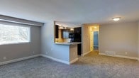 This single-floor residential unit offers a thoughtfully designed open-concept layout that seamlessly connects the kitchen and living areas. The kitchen is a standout feature, showcasing dark espresso-toned cabinetry that extends to the upper walls, complemented by black appliances and a light-toned countertop. A peninsula-style breakfast bar divides the kitchen from the living space, providing both additional counter space and a casual dining option. Track lighting overhead illuminates the kitchen, enhancing its functional appeal. The living area is finished with plush, neutral-toned carpeting that adds warmth and comfort underfoot. Neutral gray walls throughout create a clean, cohesive aesthetic that lends itself well to a variety of décor styles. A hallway extends from the main living space, leading to additional rooms, including what appears to be a bedroom with its own carpeted flooring. The layout promotes a natural flow between spaces, making the unit feel connected and well-organized. Flush-mount ceiling lighting provides consistent illumination throughout the common areas and hallway. The overall ambiance is practical and inviting, with a modern color palette and efficient use of space throughout the floor plan.