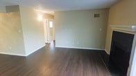 This single-floor space offers a straightforward and functional layout designed to maximize usability. The main living area is highlighted by rich, dark hardwood flooring that contrasts cleanly against the soft, neutral green-beige walls, creating a calm and understated aesthetic throughout. White baseboards provide a crisp border along the floors, adding a touch of polish to the overall finish.

The space is well-served by a ceiling-mounted HVAC vent, ensuring consistent climate control throughout the area. Electrical outlets are conveniently distributed along the walls, offering practical placement for everyday use. A textured ceiling adds subtle dimension overhead without drawing excess attention away from the room's clean lines.

A hallway visible from the main area connects to additional rooms, with a glimpse of natural light filtering in from what appears to be a window at the far end, suggesting good natural light accessibility deeper within the unit. The transition between spaces is seamless, maintaining the same dark hardwood flooring for visual consistency.

Overall, the space presents a neutral, move-in-ready environment with a modest yet cohesive design. Its clean finishes, practical layout, and warm flooring tones make it a versatile space well-suited for a variety of uses and furnishing styles.
