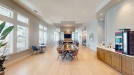 This single-floor community clubhouse is thoughtfully designed to accommodate a variety of social and recreational activities across several distinct zones. The entrance area makes an immediate impression with soaring vaulted ceilings supported by striking exposed rustic wood beam trusses, paired with bold navy blue channel-tufted seating and large windows that flood the space with natural light.

The main gathering area centers around a dramatic floor-to-ceiling textured stone fireplace wall, flanked by a mounted flat-screen television and framed by a statement sputnik chandelier overhead. Light hardwood flooring runs throughout, unifying the open layout. A generously sized dining table with patterned accent chairs anchors the central space, while smaller café-style tables offer additional casual seating near the windows.

A dedicated lounge nook features a tufted sofa, pool table, and expansive windows overlooking lush palm trees, creating a bright, resort-inspired atmosphere. A quiet seating alcove with two linen accent chairs and transom windows provides a more intimate retreat. The leasing or reception area includes semi-private booth seating finished in deep navy velvet with gold accent furnishings and curated wall art, completing this well-appointed and versatile community space.