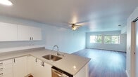 This single-floor residence is designed with a clean, modern aesthetic that emphasizes an open and functional layout. The kitchen and living area flow seamlessly together in an open-concept arrangement, creating a spacious and connected feel throughout. The kitchen is well-appointed with stainless steel appliances, including a refrigerator, electric range, and dishwasher. White flat-panel cabinetry lines the walls and provides generous storage, while neutral-toned countertops extend across an L-shaped peninsula that doubles as a breakfast bar and natural divider between the kitchen and living space. Wood-look flooring runs consistently throughout the main living areas and into the bathroom, contributing to a cohesive and contemporary appearance. A ceiling fan centered in the main living area offers both practical ventilation and ambient lighting. The living room is generously proportioned and benefits from a large window that frames lush greenery outside, inviting natural light deep into the space. The bathroom features the same warm wood-look flooring, clean white walls, and chrome fixtures, maintaining the consistent modern tone found throughout. Overall, the unit presents a bright, neutral palette with durable finishes suited for everyday living.