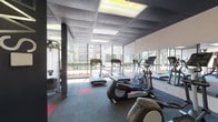 This fitness center offers a comprehensive workout environment designed to accommodate a wide range of exercise needs. The space is organized into distinct functional zones, providing ample room for both cardio training and strength work. Large floor-to-ceiling windows line the perimeter walls, bringing in abundant natural light and offering views of the surrounding neighborhood, creating an open and energizing atmosphere. The flooring consists of durable commercial carpet tiles in a gray and red accent pattern, with designated red mat areas marking specific workout zones.

The cardio section is outfitted with multiple treadmills and elliptical trainers from Life Fitness, positioned along the windowed walls to maximize natural light during use. Full-length mirrors are mounted along interior walls, visually expanding the space and allowing users to monitor their form. The strength training area features a Synrgy360 functional training system in a bold red frame, a cable machine, a full dumbbell and kettlebell rack, stability balls, and a barbell with weight plates. A water station is conveniently positioned within the space. Recessed panel lighting in the drop ceiling ensures consistent illumination throughout. The overall design is modern, clean, and purposefully laid out to support a complete fitness routine.