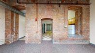 This distinctive single-floor space masterfully combines historic industrial character with modern-day functionality. The centerpiece of the main living area is a dramatic exposed brick wall featuring a striking arched passageway, which connects the open carpeted living space to the rest of the unit. Soaring wood-planked ceilings with visible beams and exposed ductwork lend an authentic loft-like aesthetic, while a pendant globe light fixture adds a warm, classic touch.

Large multi-pane windows along the painted brick perimeter walls flood the living area with abundant natural light, further enhancing the sense of openness and volume. The kitchen is well-appointed with warm wood cabinetry, dark countertops, a checkered tile floor, and a full suite of appliances including a gas range, dishwasher, and refrigerator — all framed by exposed brick that continues the building's industrial character throughout.

A dedicated corridor connects the living and kitchen areas to the bathroom and additional rooms. The bathroom is cleanly finished with white tile, a tub-shower combination, mosaic tile flooring, and a wood-framed vanity. Throughout the space, the thoughtful juxtaposition of raw historic materials and contemporary finishes creates a uniquely compelling and livable environment.