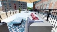 This rooftop patio is an expansive outdoor amenity space situated atop a multi-story building, accessible via an interior corridor marked with directional signage. The patio is enclosed by black metal railings that provide safety while preserving unobstructed panoramic views of the surrounding urban landscape, including notable brick-façade buildings with large industrial-style windows and active city streets below.

The space is organized into several distinct functional zones. A lounge area features a sectional sofa with patterned cushions and bold red accent pillows, arranged around a woven coffee table set atop a blue geometric-print area rug, all shaded by a large cantilever umbrella. A separate dining area includes a wicker table with matching chairs, also positioned beneath an umbrella for sun protection. Additional seating options include individual chairs positioned along the perimeter railing, ideal for taking in street-level views.

Potted evergreen topiaries and planters are distributed throughout the patio, introducing greenery and structure to the open space. The concrete deck surface is clean and well-maintained, contributing to a polished, contemporary aesthetic. The patio benefits from abundant natural light and open sky exposure, creating an inviting atmosphere for outdoor relaxation or social gatherings within an urban environment.