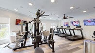 Located at 1500 Laurel Crossing Pkwy NE in Buford, GA, this single-floor fitness and wellness facility is thoughtfully designed to support a well-rounded active lifestyle. The space is divided into distinct functional areas, each finished with light wood-look flooring, crisp white trim, and neutral tones that create a clean, welcoming atmosphere throughout.

The main fitness room is generously sized and outfitted with a full complement of equipment, including treadmills, elliptical machines, a multi-station cable machine, a weight bench, and a dumbbell rack. Large windows allow natural light to pour in, while ceiling fans and wall-mounted televisions enhance comfort during workouts. Full-length mirrors along one wall amplify the sense of space.

Adjacent to the gym is a dedicated exercise studio, ideal for yoga, stretching, or group classes. This room features serene blue accent walls, warm wood-look floors, yoga mats, and a large wall-mounted mirror with a barre. A mounted television and vibrant wall art contribute to an energizing yet calming environment. A built-in kitchenette with white cabinetry and granite-style countertops adds convenience to the studio space.

A motivational word-art mural anchors one wall of the gym area, reinforcing the facility's wellness-focused ethos. The overall design balances functionality with aesthetic appeal, making it an inviting destination for fitness and well-being.