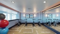 This single-level spin studio is designed to provide a focused and energizing workout environment. The room is outfitted with a row of stationary cycling bikes arranged in a forward-facing layout, offering ample space between each unit for comfortable use. A full-length mirror spans one wall, visually expanding the space and allowing cyclists to monitor their form during workouts. The walls are painted in a soft, calming blue tone, contributing to a clean and refreshing atmosphere well-suited for exercise. Two framed nautical-themed artworks are centered on the main wall, adding a subtle decorative touch that complements the cool color palette. Wall-mounted sconce lighting provides warm, even illumination throughout the room, supplemented by recessed ceiling lights. Natural light filters in through windows along one side of the room, brightening the space. The flooring consists of patterned carpet tiles in neutral tan and gold tones, offering both cushioning and durability underfoot. A textured ceiling and visible HVAC vents indicate thoughtful climate control, ensuring comfort during intense cycling sessions.