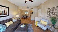 This single-floor home offers an open, flowing layout that connects the main living areas with ease. The living room is a comfortable and well-appointed space, featuring a ceiling fan, neutral beige walls, and warm hardwood-style flooring that extends throughout the home. The room accommodates a generous seating arrangement, and a half-wall partition subtly defines the entry area without disrupting the open feel.

Transitioning naturally from the living room, the dining area is highlighted by a tray ceiling detail and a pendant light fixture that adds a touch of character. A round dining table with dark chairs creates an intimate setting, framed by a window that draws in natural light.

Adjacent to the dining space, the kitchen is accessible through an open pass-through, giving the home a connected and social atmosphere. It features light wood cabinetry, a tile backsplash, and modern appliances, providing both functionality and style.

Throughout the home, the consistent hardwood-style flooring, soft neutral tones, and thoughtful layout create a cohesive and inviting ambiance. The open-concept design maximizes the sense of space, making this a practical and comfortable single-floor residence.