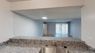 This single-floor space is designed with a clean, modern aesthetic that emphasizes an open and airy feel throughout. The living area is generously proportioned, featuring light-toned wood-style flooring that flows seamlessly throughout the unit, neutral gray walls, and a textured ceiling. A full-width sliding glass door fitted with vertical blinds anchors one wall of the living area, allowing abundant natural light to fill the space while providing access to the outdoors.

The kitchen is positioned adjacent to the living area in an open-concept arrangement, separated by a peninsula-style counter that doubles as a casual dining or prep surface. The kitchen is equipped with granite countertops featuring a subtle gray and white pattern, gray cabinetry, stainless steel appliances including a refrigerator and range, and an overhead range hood. A stainless steel sink with a standard faucet is integrated into the countertop, offering a direct sightline into the living space.

The bathroom continues the cohesive design language, featuring matching granite countertops, gray cabinetry with chrome hardware, and white fixtures. Chrome towel bars and accessory hardware add a polished finishing touch. The overall palette of whites, grays, and natural wood tones lends the space a fresh, contemporary character throughout.