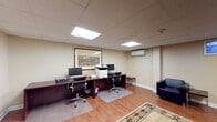This single-floor commercial office space is designed to support a range of professional functions within an open, well-organized layout. The space is finished with warm hardwood flooring throughout, white-trimmed walls in a neutral beige tone, and a drop tile ceiling equipped with recessed fluorescent panel lighting that provides consistent, even illumination across the entire area.

The workstation area is anchored by a dark wood L-shaped desk configuration accommodating two computer workstations, complete with ergonomic office chairs, a multifunction printer, and chair mats. A large framed aerial map serves as a focal point on the wall behind the desks, lending a professional character to the work zone.

Adjacent to the workstation area, an open meeting and reception zone features a round conference table with a task chair alongside multiple black leather armchairs and a low side table, centered by a decorative area rug with a geometric border pattern. Wall-mounted artwork adds visual interest throughout.

Climate control is provided by a wall-mounted mini-split HVAC unit, and a small high window with a decorative blue panel covering allows for subtle natural light. A black-framed glass door connects to an adjacent corridor or entry area, enhancing accessibility and flow within the space.