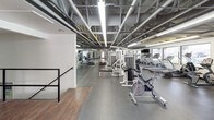 This single-floor fitness center offers a comprehensive workout environment designed to accommodate a wide range of exercise needs. The facility features an exposed structural ceiling with steel beams and industrial fluorescent lighting, lending the space a utilitarian yet functional aesthetic throughout.

The cardio area is extensively equipped with multiple treadmills, elliptical machines, stair climbers, and upright stationary bikes arranged along windowed walls. Large windows line the perimeter, allowing natural light to fill the space and providing outdoor views of surrounding greenery. Mounted flat-screen televisions are positioned throughout for entertainment during workouts.

The strength training section includes a multi-station cable and weight machine setup, accommodating a variety of resistance exercises. A dedicated free weights area features a full dumbbell rack with an organized selection of weights. Mirrored wall panels in the strength zone enhance the sense of space and allow users to monitor their form.

A separate functional training area includes a heavy bag on a freestanding stand, alongside a wood-floor zone suitable for stretching or bodyweight exercises. The facility is accessed through glass double doors and features durable rubber flooring throughout the main workout areas, ensuring safety and comfort during exercise.