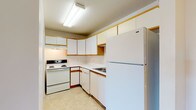This single-floor residence offers a practical and comfortable layout designed for everyday living. The kitchen is efficiently arranged in a U-shaped configuration, featuring white cabinetry with warm wood-tone trim accents, white appliances including a coil electric range with overhead ventilation, a dishwasher, and a full-size refrigerator. Light tile flooring and a fluorescent overhead fixture keep the space bright and functional. Adjacent to the kitchen is a dedicated dining area, also finished with tile flooring, providing a seamless transition between cooking and eating spaces. The living room is generously sized, finished with neutral carpet and bathed in natural light from a window, creating a welcoming atmosphere. The bedrooms continue the warm, carpeted aesthetic, each featuring windows with horizontal blinds that allow for natural light and outdoor views. The full bathroom is clean and straightforward, offering a tiled tub-shower combination, toilet, and a vanity with ample counter space, all set on neutral tile flooring. A standout feature of this residence is the dedicated laundry room, which houses a full-size washer and dryer alongside a water heater, adding a layer of convenience seldom found in similar-sized homes. Throughout, the interiors are finished in crisp white tones with clean lines.