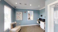 This single-floor business center is designed to provide a functional yet inviting environment for work and productivity. The space is finished with soft blue walls, crisp white crown molding, and neutral carpet tiles, creating a calm and professional ambiance throughout.

A built-in wooden workstation lines one wall, equipped with a desktop computer, printer, and ample drawer storage, offering a dedicated and organized workspace. Recessed ceiling lights provide bright, even illumination across the room, ensuring a well-lit environment suitable for focused work. A framed interior window brings in additional light from an adjoining area, adding an open, connected feel to the space.

A comfortable upholstered chaise lounge is positioned near the workstation area, offering a relaxed seating option for visitors or those taking a break. Decorative wall art featuring a colorful umbrella motif adds a touch of personality and warmth to the otherwise clean and understated interior. The space also features a security camera, ensuring a safe and monitored environment. Overall, this business center balances practicality with a welcoming aesthetic, making it a well-rounded facility for professional use.