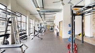 Located at 1111 Hawthorne Lane in Charlotte's Plaza Midwood neighborhood, this fitness facility offers a thoughtfully designed, two-room exercise space spread across a single floor. The design masterfully blends historic character with modern functionality, featuring striking exposed brick and stone accent walls alongside crisp white surfaces and soaring ceilings with visible ductwork — a hallmark of industrial-chic architecture.

The main exercise room is bathed in natural light streaming through large floor-to-ceiling windows that frame views of the surrounding brick building exterior and an outdoor pool area. This generously proportioned space is outfitted with a full row of treadmills, cable machines, strength equipment, a smith machine, flat bench, dumbbell rack, and a heavy punching bag — catering to a wide range of fitness routines. Pendant lighting and exposed ceiling conduits add to the loft-inspired aesthetic.

The second exercise room provides a dedicated functional training zone, featuring a multi-station rig with TRX suspension trainers and battle ropes, a floor-to-ceiling mirror wall, spin bikes, stability balls, medicine balls, and a power tower. Durable rubber flooring runs throughout both rooms, ensuring comfort and safety. Together, the two rooms form a comprehensive, well-appointed fitness destination with character and substance.