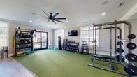 This single-story fitness center in Greensboro, NC is thoughtfully designed to accommodate a wide range of workout styles within a clean, well-appointed layout. The space is divided into two primary zones: a strength and functional training area and a dedicated cardio room, each finished with attention to both form and function.

The strength zone features green turf flooring, providing a durable, performance-friendly surface ideal for functional movement and weightlifting. Equipment includes a multi-station power rack, a full dumbbell rack with an extensive weight selection, plyo boxes, TRX suspension trainers, BOSU balance trainers, medicine balls, and a cable pulley machine. Large windows allow natural light to fill the room, complemented by recessed lighting and a large ceiling fan overhead. Wall-mounted mirrors expand the visual space and allow users to monitor their form during lifts.

The cardio area transitions to wood-look hard flooring and houses a row of treadmills, ellipticals, and a stationary bike, all positioned facing a wall-mounted flat-screen television. A matching ceiling fan and additional windows maintain comfort and airflow throughout. Double glass doors open to an outdoor view, connecting the interior to the surrounding greenery. Crown molding and neutral gray walls lend a polished, community-grade aesthetic throughout the entire facility.
