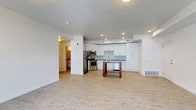 This single-floor space offers a clean, modern layout centered around an open-concept kitchen and living area. The kitchen is well-appointed with white shaker-style cabinetry that extends to the ceiling, a blue-gray subway tile backsplash, and a full suite of stainless steel appliances including a refrigerator, electric range with microwave above, and dishwasher. A freestanding wooden kitchen island provides additional workspace and casual dining potential. Recessed lighting illuminates the kitchen area, complementing the natural light that fills the space. Light hardwood-style flooring runs throughout, creating a cohesive and airy aesthetic. The open living area flows seamlessly from the kitchen, offering generous room for flexible furnishing arrangements. A textured ceiling adds subtle character to the otherwise crisp, white interior. A hallway visible from the main area leads to additional rooms, suggesting a practical and well-organized floor plan. Floor vents and recessed lighting underscore the functional infrastructure of the space. Overall, the unit presents a bright, neutral palette that provides an excellent canvas for a variety of interior styles.