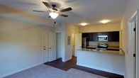 This single-floor residence offers an open-concept design that connects the living area and kitchen in a comfortable, functional layout. The living space features plush carpet underfoot and is topped with a classic ceiling fan with integrated lighting, providing both comfort and airflow throughout the room. Warm yellow-toned walls lend a welcoming ambiance to the interior.

The kitchen is a standout feature, showcasing dark cabinetry that contrasts elegantly with light granite countertops. A peninsula-style island extends the counter space, offering room for casual dining or meal preparation. Stainless steel appliances — including a refrigerator, electric range, and built-in microwave — complete the modern kitchen setup. Rich hardwood-style flooring flows through the kitchen and dining area, creating a clean transition from the carpeted living space.

Multiple doors lead to additional rooms and closet storage, hinting at a practical and well-organized floor plan. Recessed and flush-mount ceiling lights ensure the space is well-lit throughout. The overall design balances everyday functionality with a clean, contemporary aesthetic, making it a highly livable and inviting single-floor home.