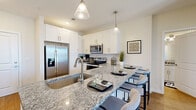 This single-floor residence is thoughtfully designed with a contemporary aesthetic and a cohesive neutral color palette throughout. The kitchen serves as the centerpiece of the living space, showcasing an expansive granite-topped island with seating for four, ideal for casual dining and entertaining. White shaker-style cabinetry lines the walls, complemented by stainless steel appliances including a French door refrigerator, range, and microwave. Pendant lighting above the island adds a polished finishing touch, while wood-style flooring flows seamlessly through the main living areas.

The bedrooms offer comfortable retreats, each featuring large windows dressed with blinds and floor-length curtains that allow for natural light and outdoor views. The primary bedroom provides generous space, while a secondary bedroom includes a built-in desk area, making it well-suited as a home office or guest room.

The bathrooms are finished with neutral tile surrounds and soaking tubs, delivering a clean, spa-like atmosphere. The overall layout, visible in the 3D floor plan, reveals a well-organized and functional flow between rooms, making this residence both practical and visually appealing.