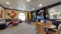 This single-floor community space is designed to accommodate a variety of activities within one open, well-appointed room. The warm yellow walls, accented by dark crown molding and trim, create an inviting atmosphere complemented by recessed lighting and a ceiling fan overhead. A textured popcorn ceiling adds dimension to the space.

A central focal point is the dark wood built-in entertainment center, which houses a flat-screen television and decorative shelving, flanked by a handsome stone fireplace beneath. This area is arranged with comfortable striped armchairs and a nearby dining table, making it suitable for both casual viewing and conversation.

Toward the far end of the room, a cozy lounge area features a brown sofa with accent pillows and a red ottoman, positioned near an arched window that allows natural light to filter in. A small dining set nearby provides an additional gathering spot. Decorative wall plates and framed artwork add character throughout.

The room is unified by a bold leopard-print carpet that runs throughout, lending a distinctive and memorable design element to this versatile, communal living space.