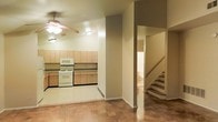 This two-story space offers a straightforward, functional layout with a clean and neutral aesthetic throughout. The main level features an open-concept design where the kitchen flows naturally into the adjacent living area, creating a cohesive and practical living environment. The kitchen is equipped with light wood cabinetry that extends along the full length of the wall, providing ample storage space. White appliances, including a gas range and built-in microwave, complement the simple, utilitarian design. The flooring on the main level consists of patterned tile that adds subtle texture to the space, while neutral beige walls maintain a bright and airy atmosphere. A ceiling fan with an integrated light fixture is centrally positioned, offering both ventilation and overhead lighting to the combined kitchen and living area. A structural column and open archway define the transition between spaces while preserving the open feel of the floor plan. A staircase with wood-toned steps and a simple railing leads to the second floor, which can be seen through the opening, suggesting additional living or sleeping quarters above. The overall ambiance is clean, understated, and move-in ready.