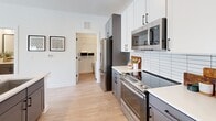 Located at 2020 Winters Eve Dr in Charlotte, NC, this single-floor studio offers a thoughtfully designed open-concept layout that seamlessly blends living and sleeping areas. Light wood-style flooring runs throughout, complementing the bright white walls and creating a clean, cohesive aesthetic.

The living area is bathed in natural light from multiple large windows dressed with soft teal curtains and plantation-style blinds. A ceiling fan overhead adds both comfort and style to the space.

The kitchen is a standout feature, boasting two-tone cabinetry—white upper cabinets paired with darker lower units—along with stainless steel appliances, a white subway tile backsplash, quartz countertops, and a peninsula island with seating. The layout provides ample prep and storage space.

The bathroom is well-appointed with gray cabinetry, a large framed mirror, matte black fixtures, a white quartz vanity top, and a tiled combination tub and shower with a curved rod. Wood-look tile flooring ties the space together with warmth and practicality.

A closet/laundry area visible from the kitchen adds functional storage. The overall ambiance of this studio is modern yet inviting, with a neutral palette and contemporary finishes throughout.