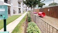 This residential community exterior showcases thoughtfully designed outdoor spaces that balance practicality with comfort. The grounds are lined with manicured green lawns, mature trees with carefully mulched bases, and neatly trimmed shrubs that add texture and color to the landscape. Smooth concrete walkways wind through the property, connecting buildings and amenity areas while providing easy pedestrian access throughout the community. A standout feature of the property is the dedicated dog park, enclosed by sturdy metal railing fencing and bordered by a tall wooden privacy fence. The dog park is equipped with playful amenities including a red tunnel and a bench, offering a safe and engaging space for pets. Posted rules signage ensures the area remains organized and enjoyable for all residents. The multi-story residential buildings feature neutral gray siding, white-trimmed windows, and private balconies with iron railings, contributing to a classic and cohesive architectural aesthetic. Exterior staircases provide access to upper-level units, maintaining an open and airy feel throughout. The overall ambiance of the community grounds is inviting and well-kept, reflecting a commitment to outdoor livability and resident convenience.
