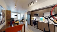 This single-floor residence offers a thoughtfully designed open-concept layout that seamlessly connects the kitchen, dining, and living areas. The kitchen is a standout feature, outfitted with dark wood cabinetry, light stone countertops, and a full suite of stainless steel appliances, including a refrigerator, electric range, and built-in microwave. Track lighting overhead provides focused illumination throughout the space, while light wood-style flooring runs continuously across the open floor plan, enhancing the sense of spaciousness.

The dining area flows naturally into a comfortable living space, which benefits from abundant natural light streaming through large sliding glass doors and windows. A ceiling fan adds to the functional design of the living room. A bedroom is visible in the background, continuing the bright, airy aesthetic of the unit.

Beyond the interior, residents have access to a beautifully landscaped outdoor amenity area featuring a resort-style swimming pool surrounded by lounge seating, a shaded pergola, and lush desert-friendly plantings. Tall palm trees frame the courtyard, enhancing the warm, sun-drenched atmosphere of the community.