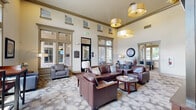 Located at 121 E Victory Road in Meridian, Idaho, this single-floor community clubhouse serves as the welcoming centerpiece of the Redtail community. The main lounge area immediately impresses with soaring double-height ceilings, large multi-pane windows, and transom windows that flood the space with natural light. Dramatic drum pendant light fixtures hang overhead, complementing the neutral palette of warm beige and taupe tones. Plush leather seating arrangements, including rich brown sofas and armchairs, are grouped around round wood coffee tables atop a patterned area rug, creating an inviting and comfortable gathering environment. A dedicated seating alcove along the windowed perimeter offers additional lounge chairs and bistro-style table seating with views of the lush exterior landscaping. The space also features a fully equipped kitchen area appointed with dark wood cabinetry, a mosaic tile backsplash, granite countertops, stainless steel appliances, and a generous island with bar seating beneath warm pendant lighting. A billiards table anchors a secondary entertainment zone, adding a recreational focal point to the layout. Textured, patterned wallcovering accents transition spaces stylishly, while decorative vases and framed artwork complete the polished, welcoming atmosphere throughout.