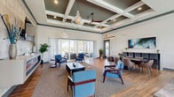 Located in Tavares, Florida, this well-appointed single-floor clubhouse is designed to serve as a refined communal gathering space. The main area is defined by its striking coffered ceilings painted in a deep sage green, complemented by elegant pendant chandeliers and ceiling fans that enhance the sense of vertical space. Large floor-to-ceiling glass doors and expansive windows flood the interior with abundant natural light, while offering views of the outdoor pool area and surrounding Florida landscaping.

The flooring throughout features a distinctive herringbone-patterned wood-look tile that adds visual texture and warmth. The open layout accommodates multiple functional zones: a casual lounge area with blue upholstered armchairs arranged around a coffee table, a dining and collaborative seating area with round tables and modern chairs, and a dedicated lounge nook anchored by a full-width linear electric fireplace set against a textured tile accent wall with a wall-mounted TV above.

The kitchen is fully equipped with dark cabinetry, granite countertops, stainless steel appliances, a herringbone mosaic tile backsplash, and a large island with bar seating — ideal for hosting events. A private seating alcove with floor-to-ceiling windows and chrome-framed chairs offers a quieter retreat. The patio extends the experience outdoors, furnished with wicker lounge seating and direct views of the pool.