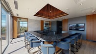 This upscale single-floor space is a masterclass in contemporary design, seamlessly blending luxury finishes with functional, multi-purpose areas. The centerpiece is a stunning wet bar and lounge featuring a large, U-shaped black marble countertop edged in rich veining, surrounded by tufted blue upholstered bar stools with brass-toned bases. Above, a striking inverted pyramid ceiling installation crafted from warm wood slats with ambient underlighting and cascading crystal elements creates a dramatic focal point. Stone-textured accent walls and warm wood paneling complete the refined aesthetic.

Adjacent to the bar, a spacious living lounge area offers deep, plush sectional seating in dark velvet tones, layered over hardwood flooring and area rugs, with a large flat-screen display and built-in shelving. A separate, intimate screening lounge provides a sunken-style seating arrangement for a more private media experience.

A bright conference room with floor-to-ceiling glass walls offers sweeping panoramic views of the city skyline and mountains, bathed in natural light. A live-edge wood conference table anchored by a sculptural pendant light serves as its focal point.

Throughout, floor-to-ceiling windows and glass sliding doors flood the space with natural light and frame breathtaking elevated city and mountain vistas, connecting the sophisticated interior to the world beyond.