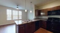 This single-floor residential unit offers a well-designed layout that balances functionality and comfort throughout. The kitchen serves as a central feature, showcasing rich dark-brown cabinetry paired with granite-style countertops and a suite of black appliances, including a range, microwave, dishwasher, and refrigerator. A double-basin stainless steel sink is positioned along the peninsula, which opens toward the dining and living areas, creating a connected and social atmosphere. Pendant lighting hangs above the kitchen, complementing the natural light that filters in through blinds-covered windows. The living and dining areas are finished with warm hardwood-style flooring that extends throughout the main living spaces, contributing to a cohesive and inviting aesthetic. A dedicated bedroom space features the same flooring and neutral wall tones, offering a clean, versatile canvas. The bathroom is well-appointed with a full tub-shower combination featuring white tile surround, a dark vanity with granite-style countertop, an undermount sink, and a large mirror. A stacked washer and dryer unit is conveniently located within the unit, adding practical everyday functionality to the overall layout.