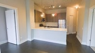 This contemporary single-floor space showcases a thoughtfully designed open-concept layout that seamlessly blends living and kitchen areas. The kitchen is a standout feature, boasting modern flat-front cabinetry in a warm taupe finish paired with a crisp white peninsula countertop that provides both prep space and a natural divider between rooms. Stainless steel appliances, including a French door refrigerator, microwave, and gas range, lend a sleek, professional aesthetic. A textured tile backsplash adds depth and visual interest to the cooking area. Overhead, a combination of track lighting and elegant pendant lights illuminate the kitchen with a warm, inviting glow. The ceiling features an exposed concrete texture that adds an industrial character to the space, contrasting beautifully with the refined finishes below. Rich, dark hardwood flooring runs throughout, grounding the space and adding warmth to the overall palette. The open layout allows natural light to flow freely, enhancing the sense of openness. Clean white walls and generous ceiling height contribute to an airy, spacious atmosphere, making this a well-appointed and stylish space suited for modern living.